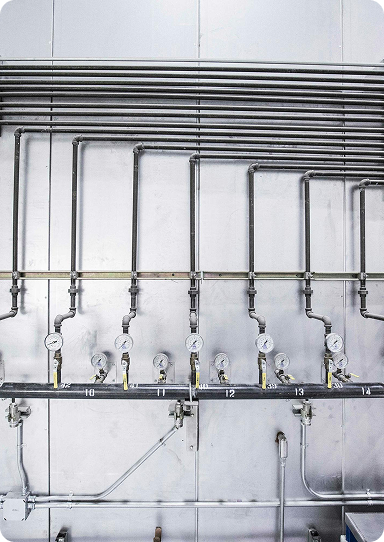 Metal pipes and pressure gauges are mounted on a wall in an industrial setting, with numbered labels beneath each gauge and valves for flow control. The background consists of stainless steel panels, reflecting the high standards maintained by professional Plumbing Services Utah County.