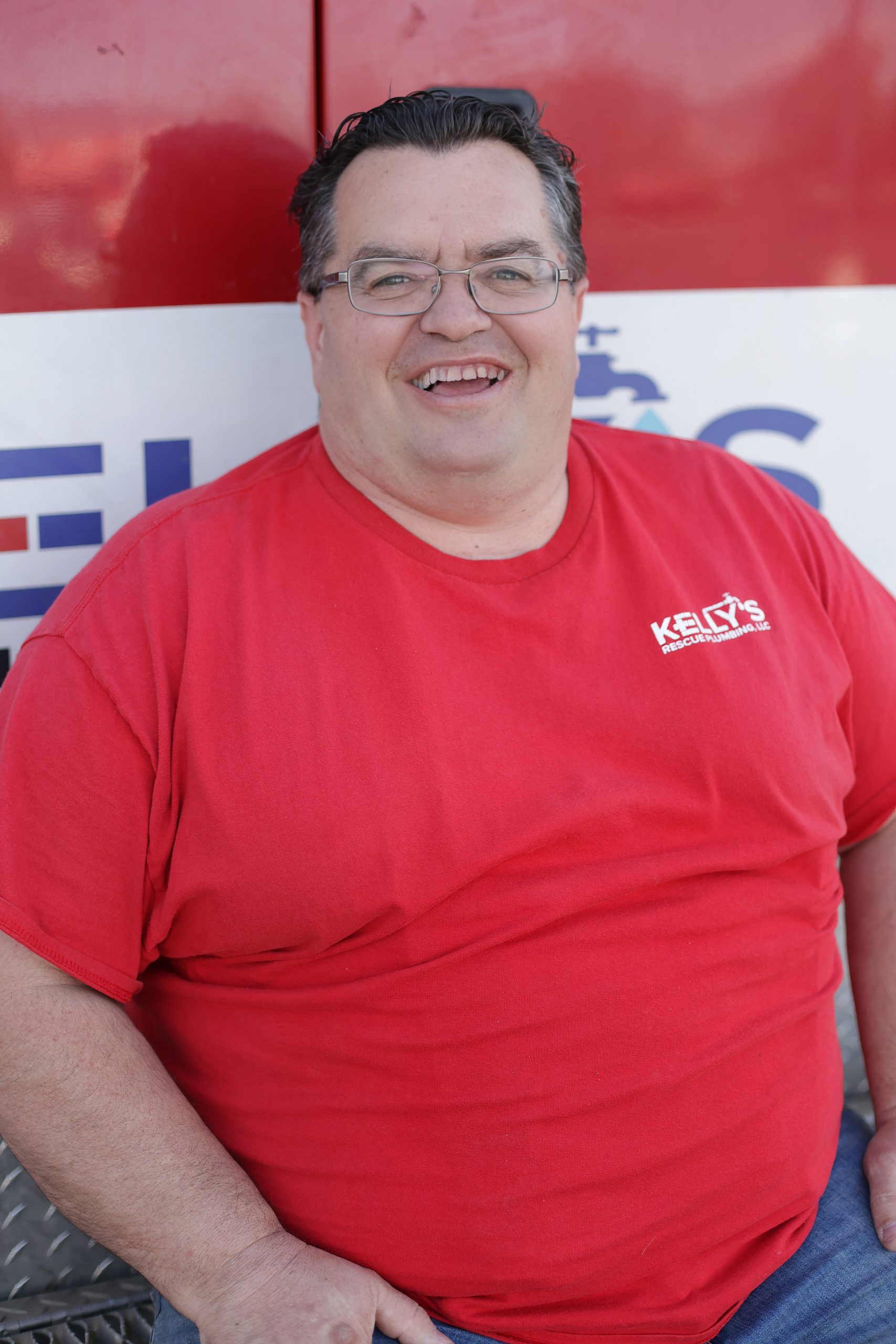 A man wearing glasses and a red “Kelly’s” t-shirt is smiling while sitting in front of a red and white background with partial text visible, representing the friendly spirit of a top plumbing team in Utah County.