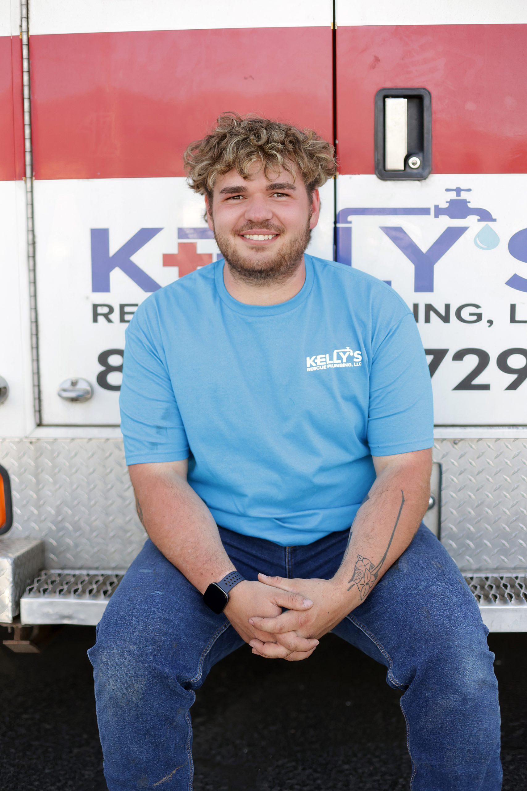 A man with curly hair, wearing a blue “KELLY’S” shirt and jeans, sits smiling in front of a service vehicle with a red stripe and partial company logo in the background, representing the trusted plumbing team serving Utah County.