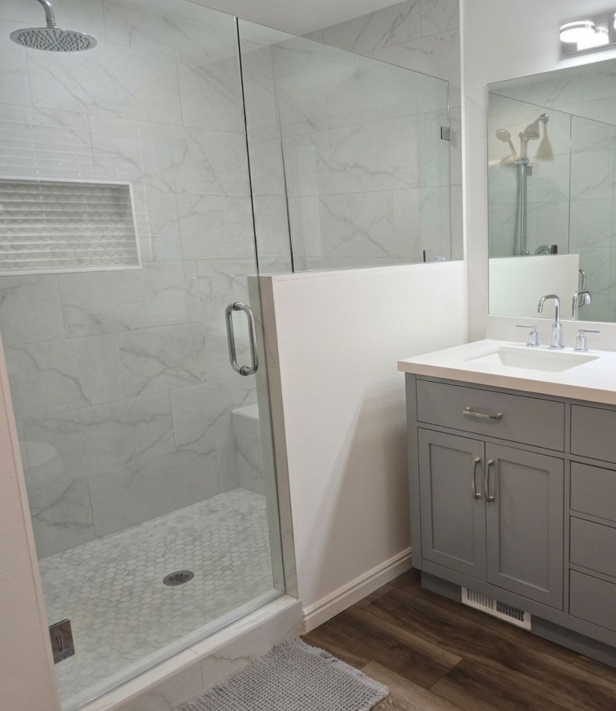 Modern bathroom with a glass-door shower, white and gray marble tiles, a gray vanity with a white sink, chrome fixtures, and a wood-style floor. A large mirror and bright lighting are above the sink—perfectly showcasing the expert workmanship from top-tier Plumbing Services Utah County.