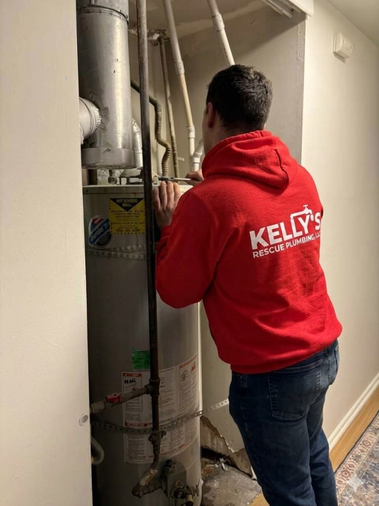 A member of the plumbing team Utah County, wearing a red “Kelly’s Rescue Plumbing” hoodie, works on a water heater in a small utility space with pipes and a white wall.