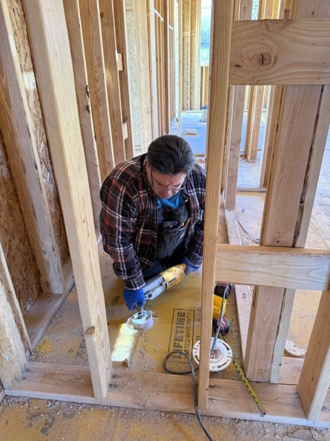 A person wearing a plaid shirt and safety glasses uses a power tool to cut a piece of wood inside a wooden-framed building under construction, where Plumbing Services Utah County may also be working to complete the project. Tools and a tape measure are on the floor nearby.