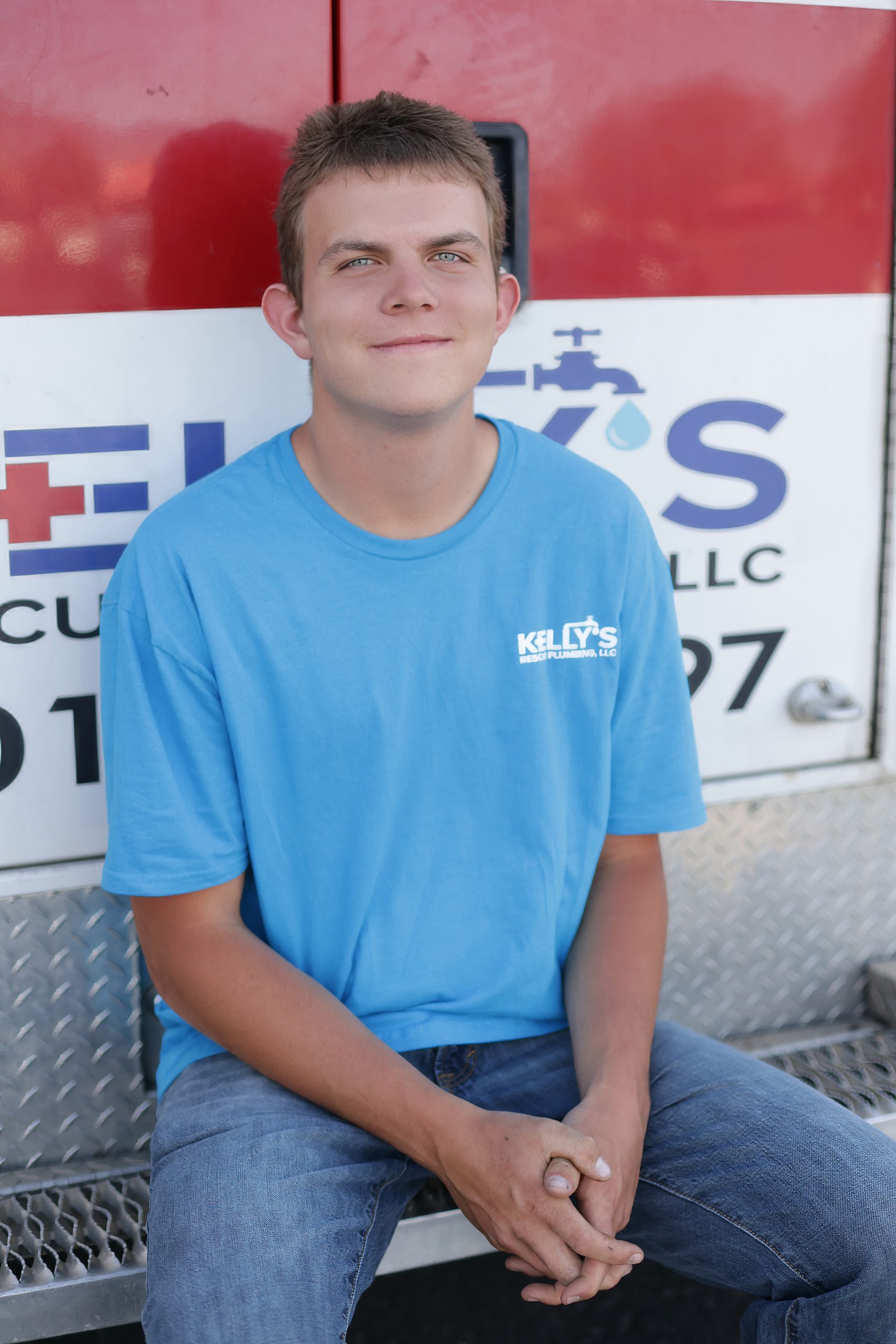 A young man in a blue “Kelly’s Plumbing” T-shirt sits on a metal step in front of a service van, smiling at the camera—a proud member of the trusted plumbing team Utah County relies on.