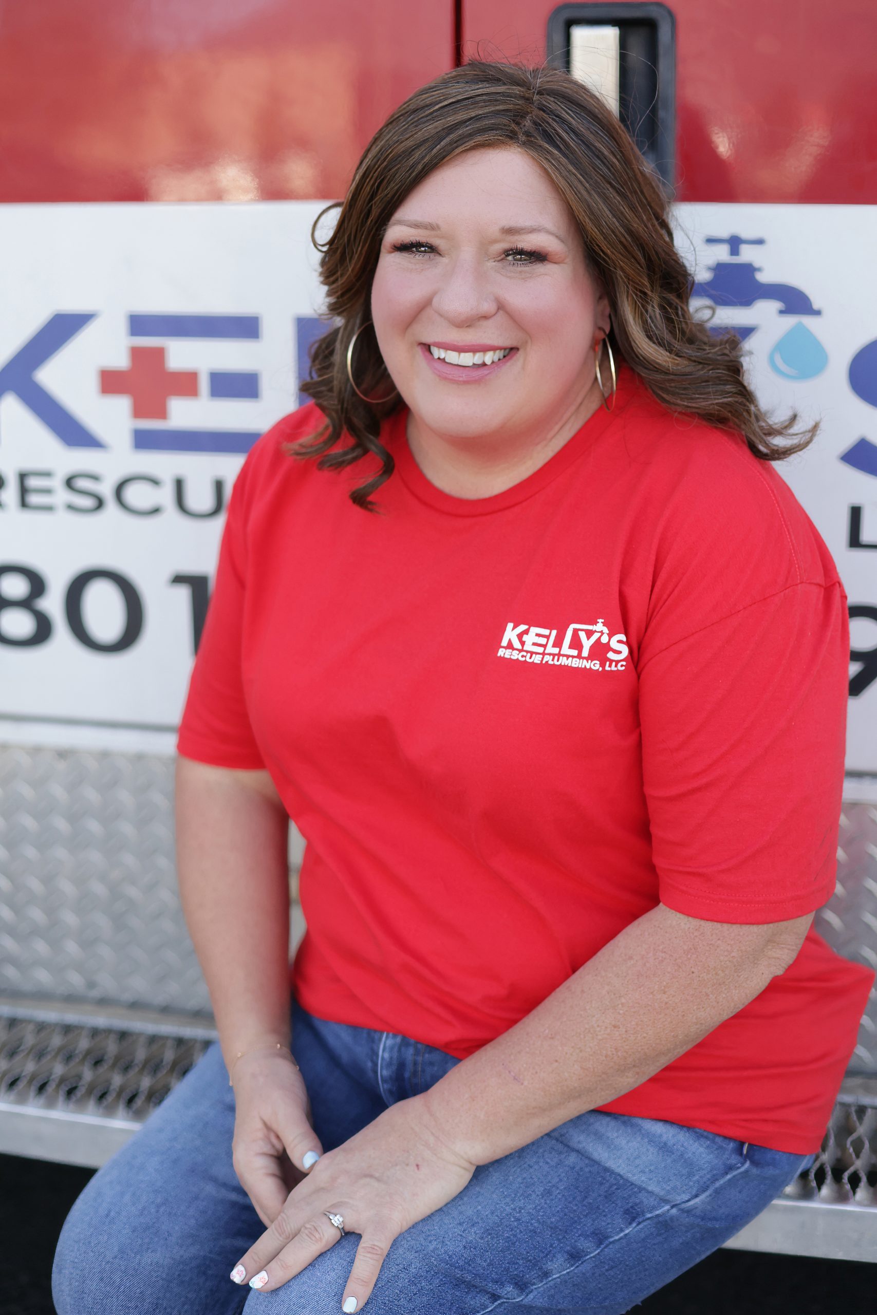 A woman with wavy brown hair wearing a red “Kelly’s Rescue Plumbing” shirt sits and smiles in front of a service vehicle with the company logo, representing the friendly plumbing team serving Utah County.