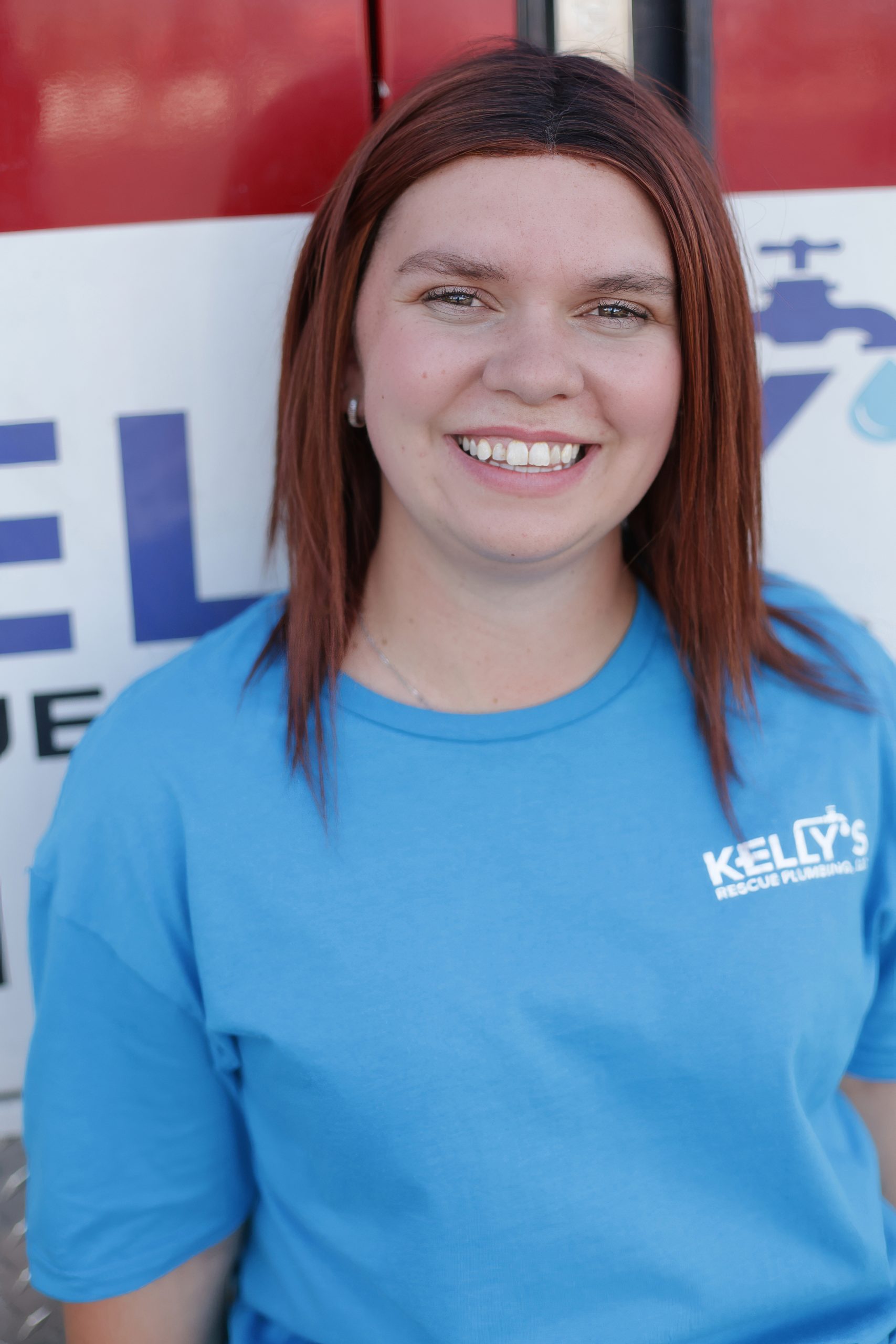 A woman with straight, reddish-brown hair in a blue “Kelly’s Rescue Plumbing” shirt smiles in front of a sign with blue letters and a faucet graphic, proudly representing the trusted plumbing team Utah County relies on.
