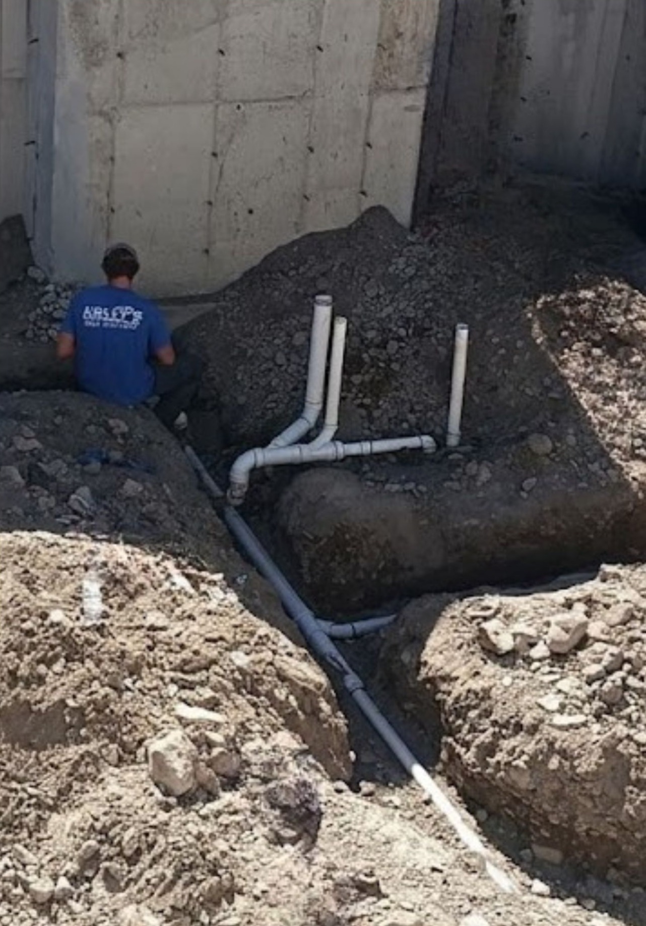A worker in a blue shirt kneels by exposed PVC plumbing pipes and dirt near a concrete wall at a construction site, demonstrating the attention to detail provided by Plumbing Services Utah County.