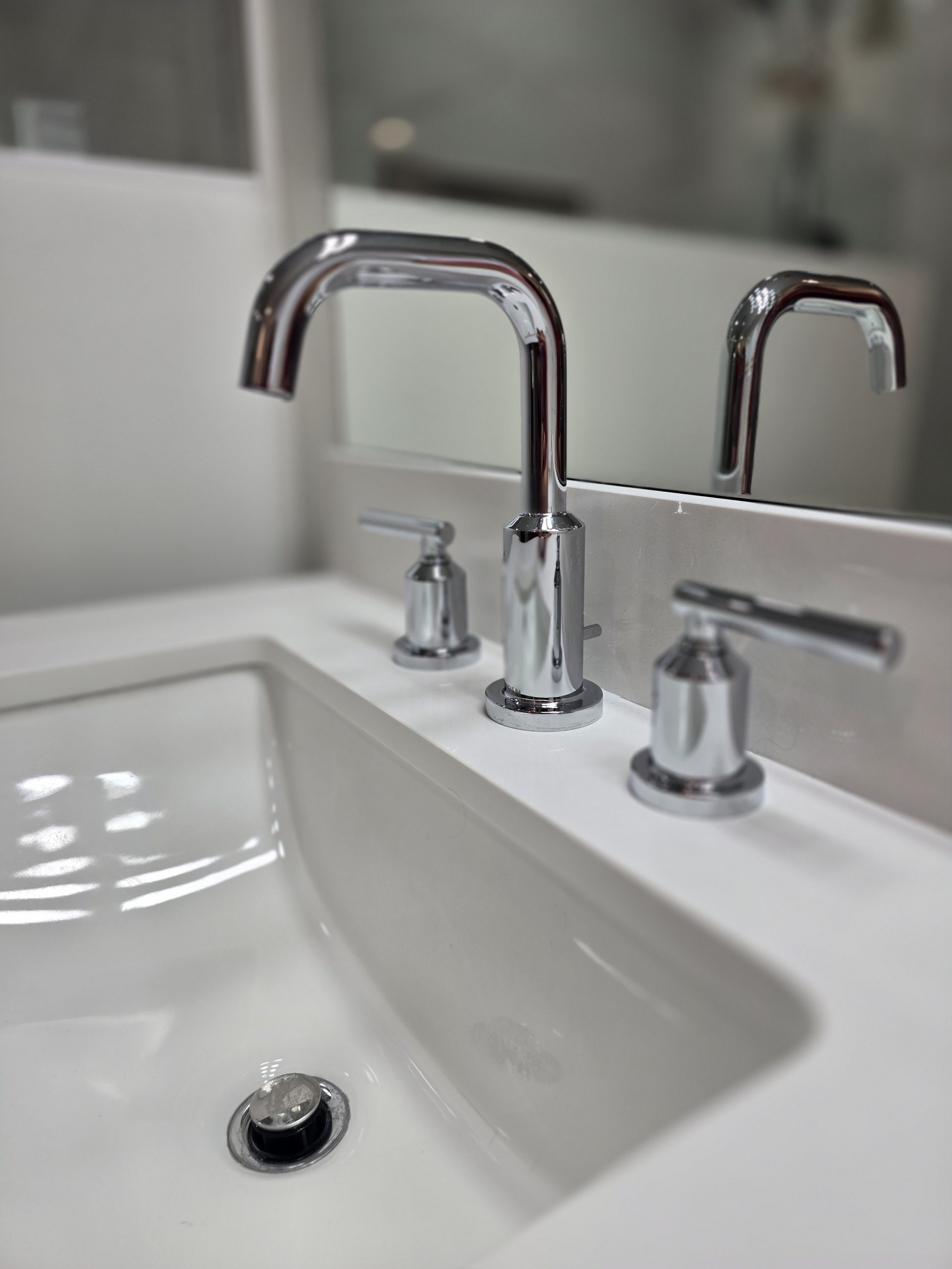 Close-up of a modern bathroom sink with a shiny chrome faucet, two handles, and a soap dispenser, all set in a white countertop. The background includes a mirror and part of a shower stall—an inviting space that reflects the high standards maintained by leading plumbing services in Utah County.
